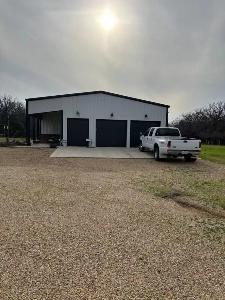 Gray metal shop building with roll-up garage doors and a pickup truck on a gravel drive